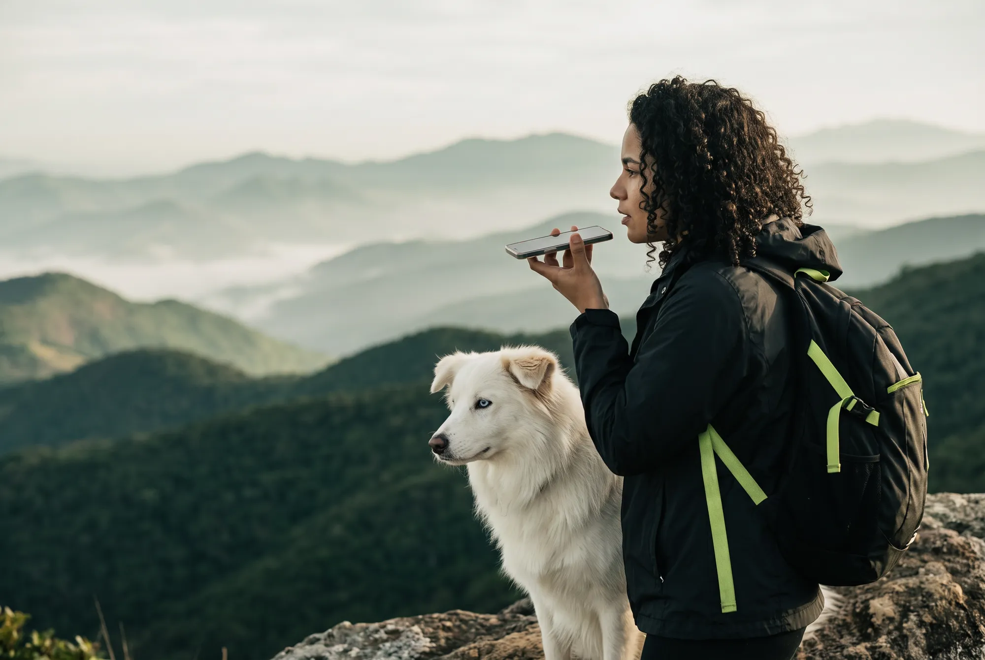 Woman at the top of a trail with the white dog beside her, recording audio on her phone, with mountains and mist behind