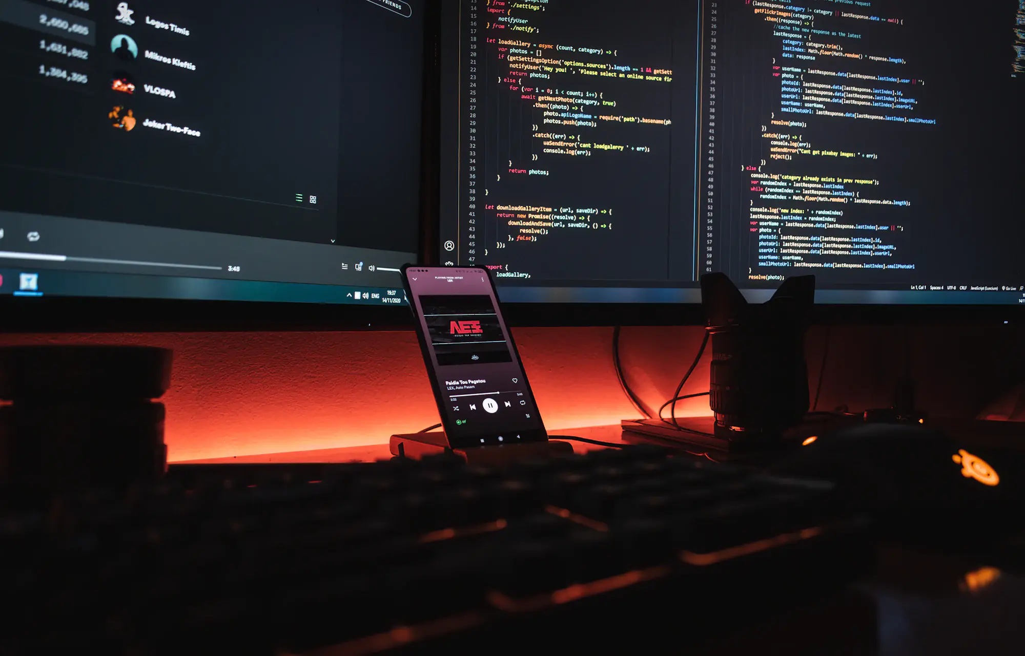 Mesa de desenvolvimento à noite, com código aberto em dois monitores e um celular apoiado na frente