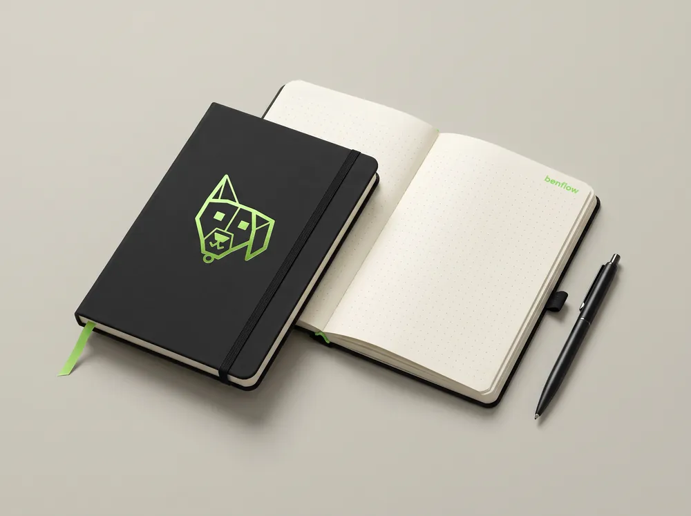 Caderno preto de capa dura com o símbolo do benflow em verde, fechado e aberto ao lado de uma caneta